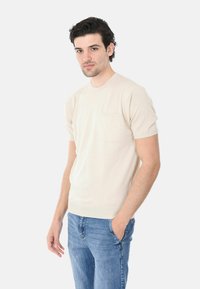 Camicia corta a maniche corte color beige chiaro con colletto rotondo e dettagli sulla tasca, abbinata a jeans blu. Tessuto liscio, design slim fit.