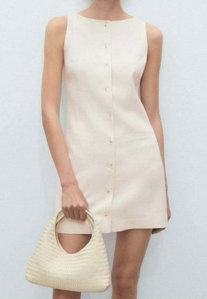 Kobieta w bezrękawowej, zapinanej na guziki beżowej mini sukience, trzymająca plecioną, kremową torebkę z okrągłymi uchwytami.