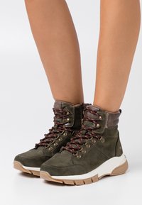 Botas de tobillo de ante verde con detalles en marrón y cordones. Presentan un cuello acolchado, suela de goma y ojales de metal para el cordón.