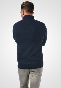 Pull en maille bleu marine avec un col montant et des poignets côtelés, porté avec un jean déchiré gris clair présentant une surface texturée.