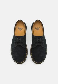 Musta mokkanahkainen Dr. Martens -kengät, joissa on keltainen tikkaus, mustat nauhat ja vaaleanruskeat nahkasisäpohjat, nähtynä ylhäältä valkoista taustaa vasten.