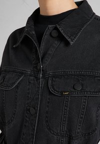 Svart denimjacka med klassisk krage, knappstängning, två bröstfickor och en liten guldfärgad logotyp på den vänstra fickan.