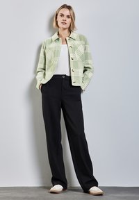 Zelená kostkovaná bunda s knoflíkovým zapínáním, bílá košile pod ní, v kombinaci s černými kalhotami a světlými botami. Jednoduchý moderní design.