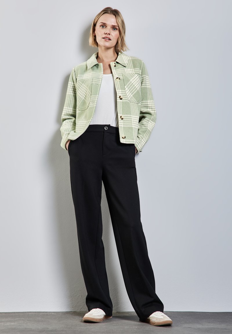 Zelená kostkovaná bunda s knoflíkovým zapínáním, bílá košile pod ní, v kombinaci s černými kalhotami a světlými botami. Jednoduchý moderní design.