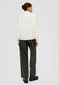 Leichte weiße Bluse mit Puffärmeln und kragenförmigem Ausschnitt, kombiniert mit dunklen, glänzenden weit geschnittenen Hosen und klobigen schwarzen Plateauschuhen.