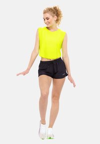 Neon yellow cropped tank top with sleeveless design, paired with black drawstring shorts. Model wears multicolored athletic shoes.