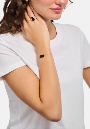 Frau trägt weißes T-Shirt mit schwarzem Ring in Form eines Gummibärchens am Finger und passendem Armband am Handgelenk vor einfarbigem Hintergrund.