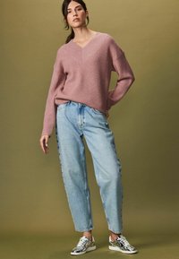 Helles pinkes geripptes Sweatshirt mit V-Ausschnitt, kombiniert mit hellblauen high-waisted Jeans mit floralen Stickereien. Silberne Sport Schuhe.