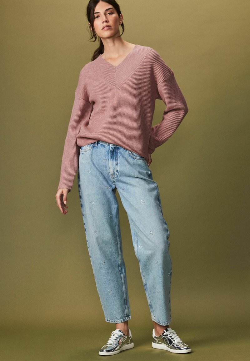 Helles pinkes geripptes Sweatshirt mit V-Ausschnitt, kombiniert mit hellblauen high-waisted Jeans mit floralen Stickereien. Silberne Sport Schuhe.