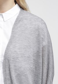 Cardigan grigio lavorato a maglia con una texture morbida, design drappeggiato e apertura frontale, indossato sopra una camicia bianca con colletto.
