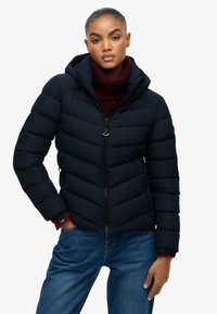 Marine blauwe pufferjas met capuchon, voorzien van een getailleerd ontwerp, horizontale naden, rits aan de voorkant en zijzakken. Draagt over een bordeaux rode sweater.