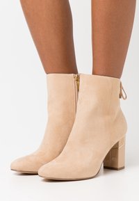 Bottines en daim beige avec un bout pointu, un détail de fermeture éclair dorée et un talon bloc texturé. Finition lisse avec un design moderne et minimaliste.