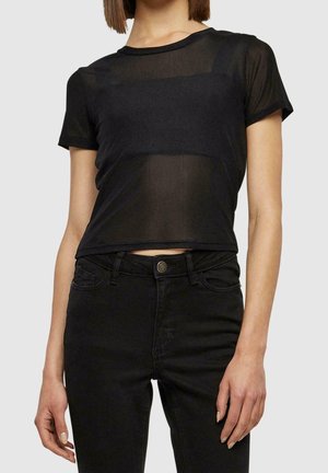 Kobieta ubrana w przezroczysty czarny krótki top z krótkim rękawem na czarnym bandeau, w wysokich czarnych dżinsach, na jednolitym tle.