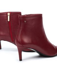 Botas de tobillo de cuero rojo con punta afilada y un tacón de 5 cm. Presentan una cremallera lateral y un pequeño logo grabado en el tobillo exterior.