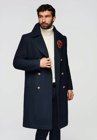 Manteau en laine bleu marine avec un design croisé, des boutons dorés, et une grande broche brodée en forme de pomme rouge sur le revers gauche.