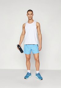 Witte atletische tanktop, lichtblauwe korte broek en blauwe sneakers. Houdt een zwarte waterfles vast. Gladde stof met een losse pasvorm.