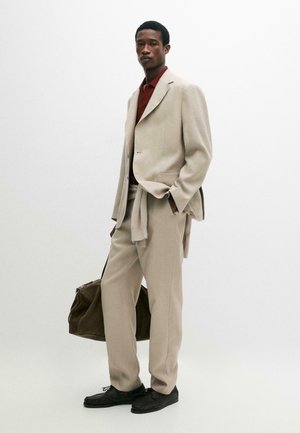 Man in een beige pak met een bordeauxrode blouse, met een bruine tas, staand tegen een witte achtergrond met één hand in de zak.