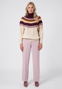 Stickad tröja med hög krage, med horisontella ränder i beige, brun, lila och gul. Kombineras med ljusrosa raka byxor.