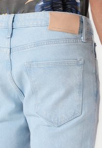 Lys blå denimbukser med en teksturert overflate, med en rektangulær lærpatch på linningen og en baklomme med dobbel søm.