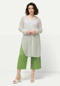 Weißes, semi-transparentes Langarmhemd mit Knopfleiste und seitlichen Schlitzen, kombiniert mit leuchtend grünen weit geschnittenen Hosen und beigen Plateausandalen.