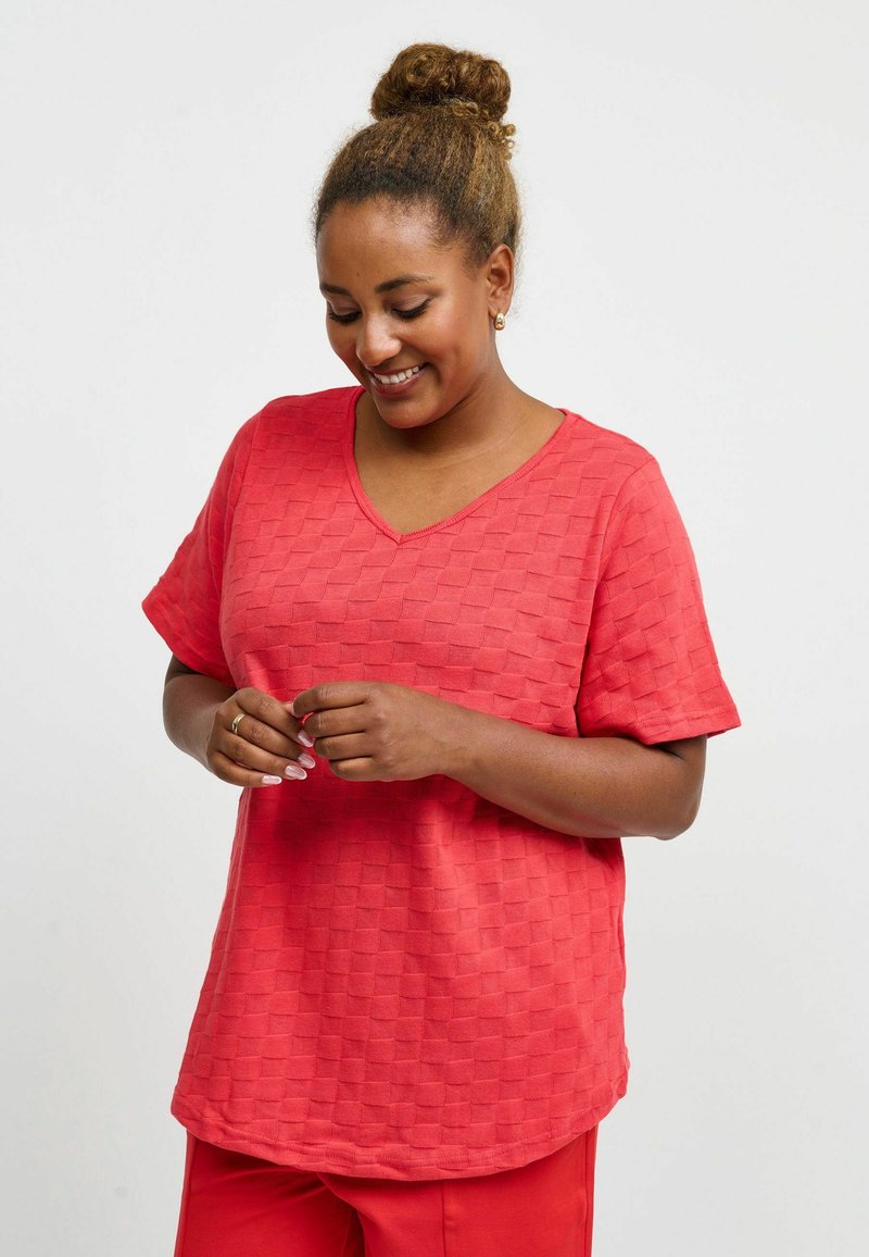 Femme souriante aux cheveux bouclés en chignon, portant un haut rouge texturé à manches courtes et un pantalon rouge assorti, regardant ses mains.