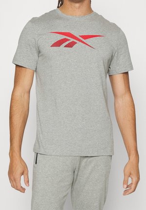 Man in een lichtgrijze t-shirt met korte mouwen en een rood gestileerd logo op de borst, gecombineerd met bijpassende lichtgrijze broek met ritszak.