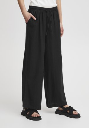 Pantaloni neri a gamba larga con vita elastica, cordoncino e tasche laterali; abbinati a sandali neri con cinturini e suola spessa.