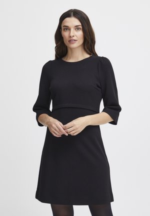 Musta mekko, jossa on lyhyet, puffimaiset hihat, teksturoitu kangas, istuva vartalo-osa ja saumatieto juuri rinnan alla. Minimalistinen muotoilu, polvimittainen.