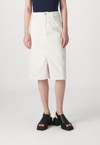 Jupe crayon en denim blanc avec couture frontale et fente, dotée d'une taille haute et d'un design à cinq poches. Associée à des sandales plateformes noires.