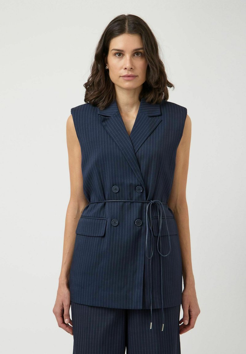Femme aux cheveux foncés mi-longs, portant un blazer bleu marine sans manches à fines rayures avec une taille nouée et un pantalon assorti.