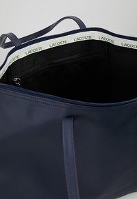 Sac cabas bleu marine en matériau durable avec fermeture éclair, comportant une bordure verte et blanche avec logo et une poche intérieure.