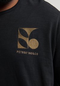 Camiseta negra con logo dorado que presenta formas abstractas y el texto "PETROL® IND&CO" en una fuente moderna, fabricada con material de algodón suave.