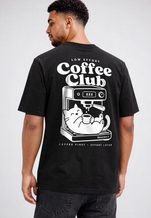 Man wearing black t-shirt with white print of a cat relaxing in a coffee machine and text "Low Effort Coffee Club" on the back.