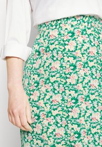 Saia floral com um fundo verde, apresentando flores rosas e brancas, cintura elástica e um tecido ligeiramente texturado.