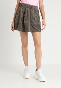 Gonna di cotone a coste marrone con pieghe, dotata di una vita elastica. Indossata con una maglietta rosa e sneakers bianche, esposta su uno sfondo neutro.