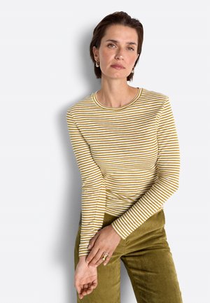 Woman with short brown hair wearing yellow and white striped long-sleeve shirt and olive green corduroy pants, standing against a light background.