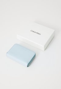 Gaiši zils ādams maciņš ar sudraba rāvējslēdzēju, izcelta logo uz priekšpuses, novietots blakus baltai kastei, kurā melnā krāsā ir izdrukāts "Calvin Klein".