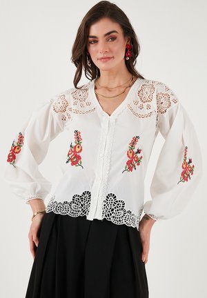 White blouse with floral embroidery in red and green, featuring lace trim, puffed sleeves, and a V-neckline. Delicate detailed accents.