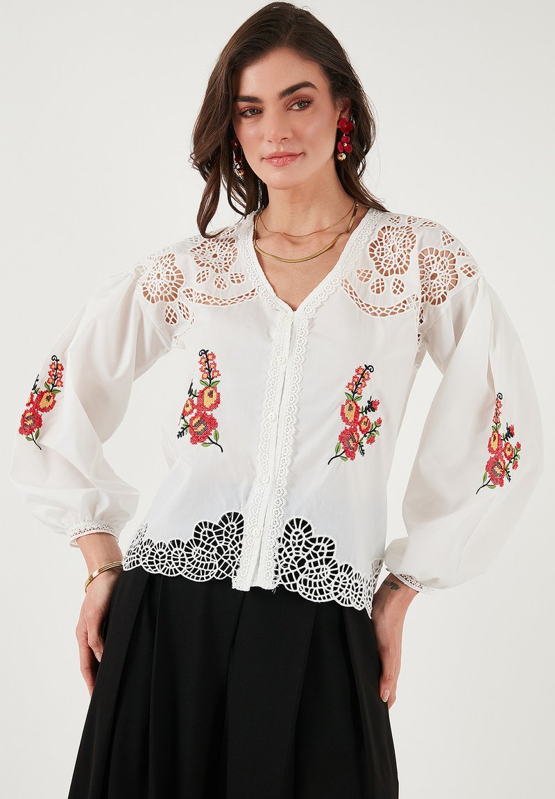 White blouse with floral embroidery in red and green, featuring lace trim, puffed sleeves, and a V-neckline. Delicate detailed accents.