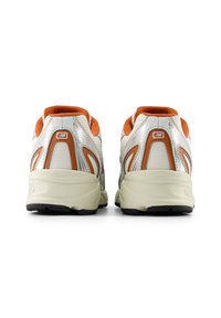 Άσπρα και ασημί αθλητικά παπούτσια με πορτοκαλί λεπτομέρειες, υφασμένο επάνω μέρος με υφή, μαλακές σόλες και μαύρες λαστιχένιες εξωτερικές σόλες, απεικονιζόμενα από πίσω.