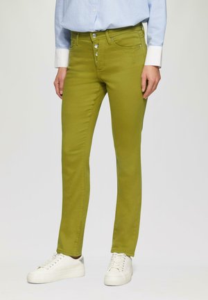 Jean skinny vert en mélange de coton avec une texture lisse, équipé d'une fermeture à cinq boutons et de poches avant. Associé à des baskets blanches.