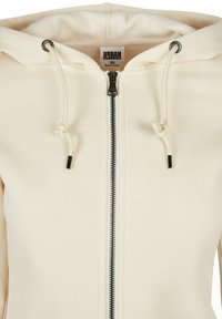Sudadera con capucha con cremallera en color crema, que presenta una textura suave, cremallera negra, cordones ajustables y una etiqueta que dice "Urban Classics."
