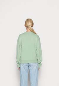 Felpa verde con vestibilità morbida, scollo rotondo e polsini a coste. Presenta un piccolo logo sul retro, vicino al collo. Abbinata a jeans azzurri.