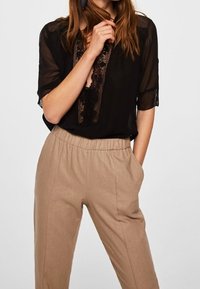 Blouse noire transparente avec détails en dentelle, associée à un pantalon beige à taille élastique, poches latérales et coupe droite.