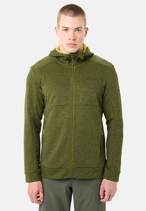 Junger Mann, der eine grüne Kapuzenjacke mit Reißverschluss, strukturierter Stoff und seitlichen Taschen trägt, vor einem schlichten hellen Hintergrund steht.