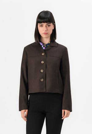 Femme portant une veste boutonnée marron foncé par-dessus une chemise colorée avec un col, accompagnée d'un pantalon noir, se tenant devant un fond blanc uni.