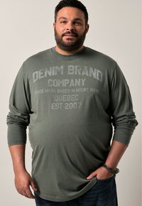 Langärmliges, olivgrünes Baumwoll-T-Shirt mit rundem Halsausschnitt und grafischem Textdruck. Getragen mit dunkelblauen Jeans.