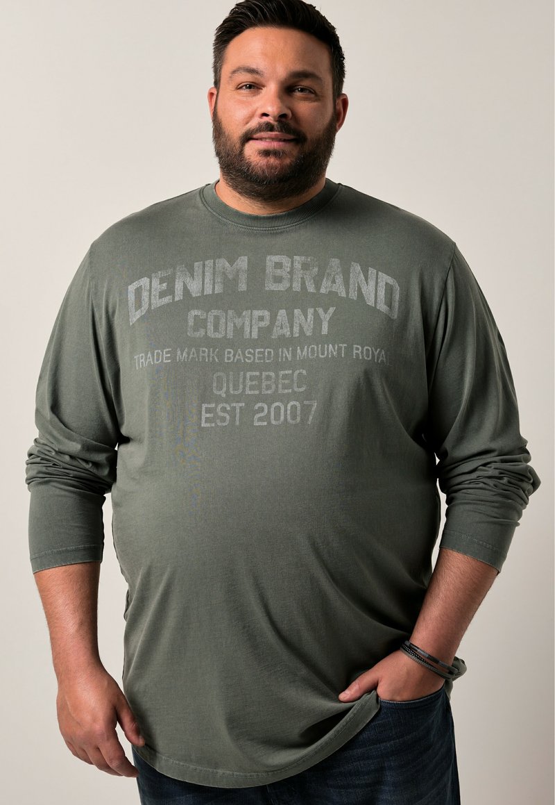 Langärmliges, olivgrünes Baumwoll-T-Shirt mit rundem Halsausschnitt und grafischem Textdruck. Getragen mit dunkelblauen Jeans.
