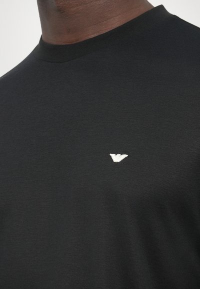 Fekete, rövid ujjú póló sima anyagból, a mellkason kis fehér hímzett logóval. Egyszerű, kerek nyakkivágás.