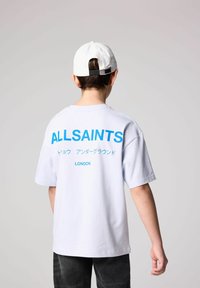 Balta medvilninė marškinė su mėlynu tekstu "ALLSAINTS" ant nugaros, su apvaliu iškirpimu ir trumpomis rankovėmis. Asmuo dėvi baltą kepurę.